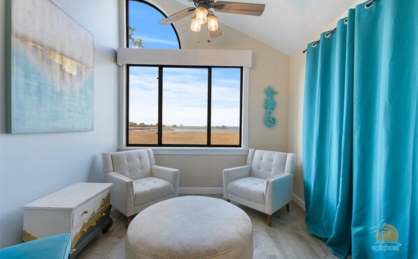A room with two white armchairs, a round ottoman, a white dresser with peeling paint, a large window with a view of a beach, and a ceiling fan.