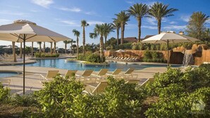 A pool area with lounge chairs and umbrellas, surrounded by palm trees and a rocky landscape. - BellaVida-Sandestin: Lakefront+Cart to Beach (Miramar Beach)