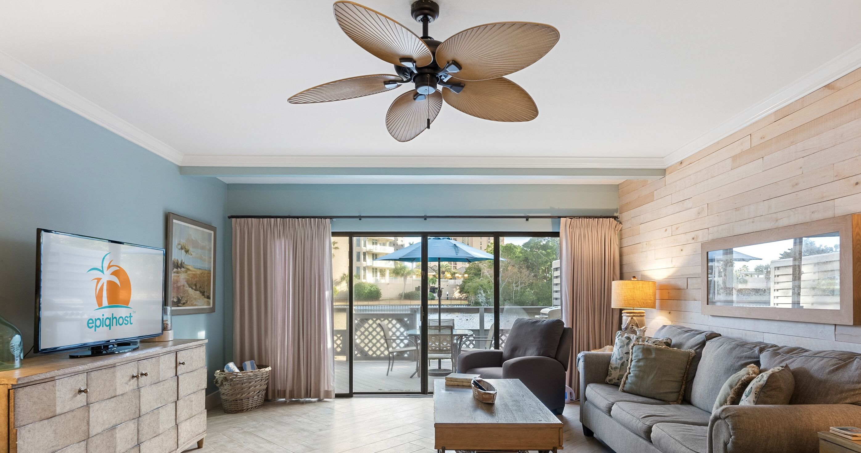 A living room with a ceiling fan, a television, a sofa, a coffee table, and a view of the outdoors through a sliding glass door.