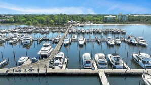 A marina with numerous boats docked at wooden piers, surrounded by greenery and buildings in the background.