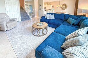 Un salon avec un canapé d’angle bleu, un fauteuil beige, une table basse ronde et un escalier menant à un niveau supérieur.