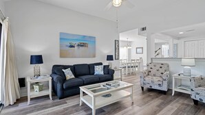 A modern living room with a sofa, coffee table, and side tables. There is a painting on the wall, a dining area in the background, and a kitchen visible through an open door. - CostaLuna-Beachwalk 2BR/2BA/Loft - Cart to Beach! (Miramar Beach)
