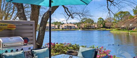 A lakeside patio with a grill, glass table, chairs, and a view of a lake and buildings.