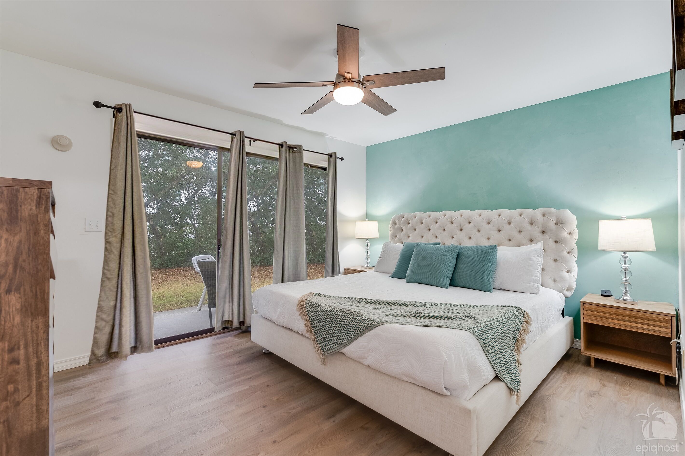 A bedroom with a large bed, a ceiling fan, a nightstand, and a view of the outdoors.