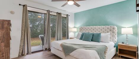 A bedroom with a large bed, a ceiling fan, a nightstand, and a view of the outdoors.