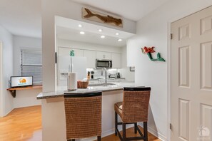 A modern kitchen with white cabinets, a stainless steel refrigerator, a microwave, and a sink. There are two wicker bar stools and a wooden shelf with a basket.