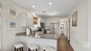 A modern kitchen with granite countertops, stainless steel appliances, and white cabinetry. There are three bar stools and a small island with a sink. - VillaIsla-3BR/2BA Villa - Steps from the Sand! (Santa Rosa Beach)