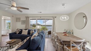 A modern living room with a sofa, dining table, and chairs. There is a ceiling fan, a large window with curtains, and a view of the outside. - VillaIsla-3BR/2BA Villa - Steps from the Sand! (Santa Rosa Beach)