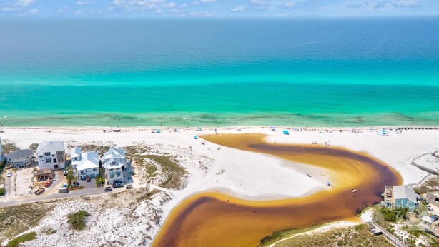 An aerial view of a beach with clear turquoise waters, a winding river of brown water, and several buildings with multiple stories and large windows. - VillaIsla-3BR/2BA Villa - Steps from the Sand! (Santa Rosa Beach)