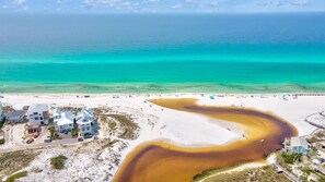 An aerial view of a beach with clear turquoise waters, a winding river of brown water, and several buildings with multiple stories and large windows. - VillaIsla-3BR/2BA Villa - Steps from the Sand! (Santa Rosa Beach)