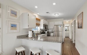 A modern kitchen with granite countertops, stainless steel appliances, and white cabinetry. There are three bar stools and a small island with a sink.