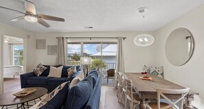 A modern living room with a sofa, dining table, and chairs. There is a ceiling fan, a large window with curtains, and a view of the outside.