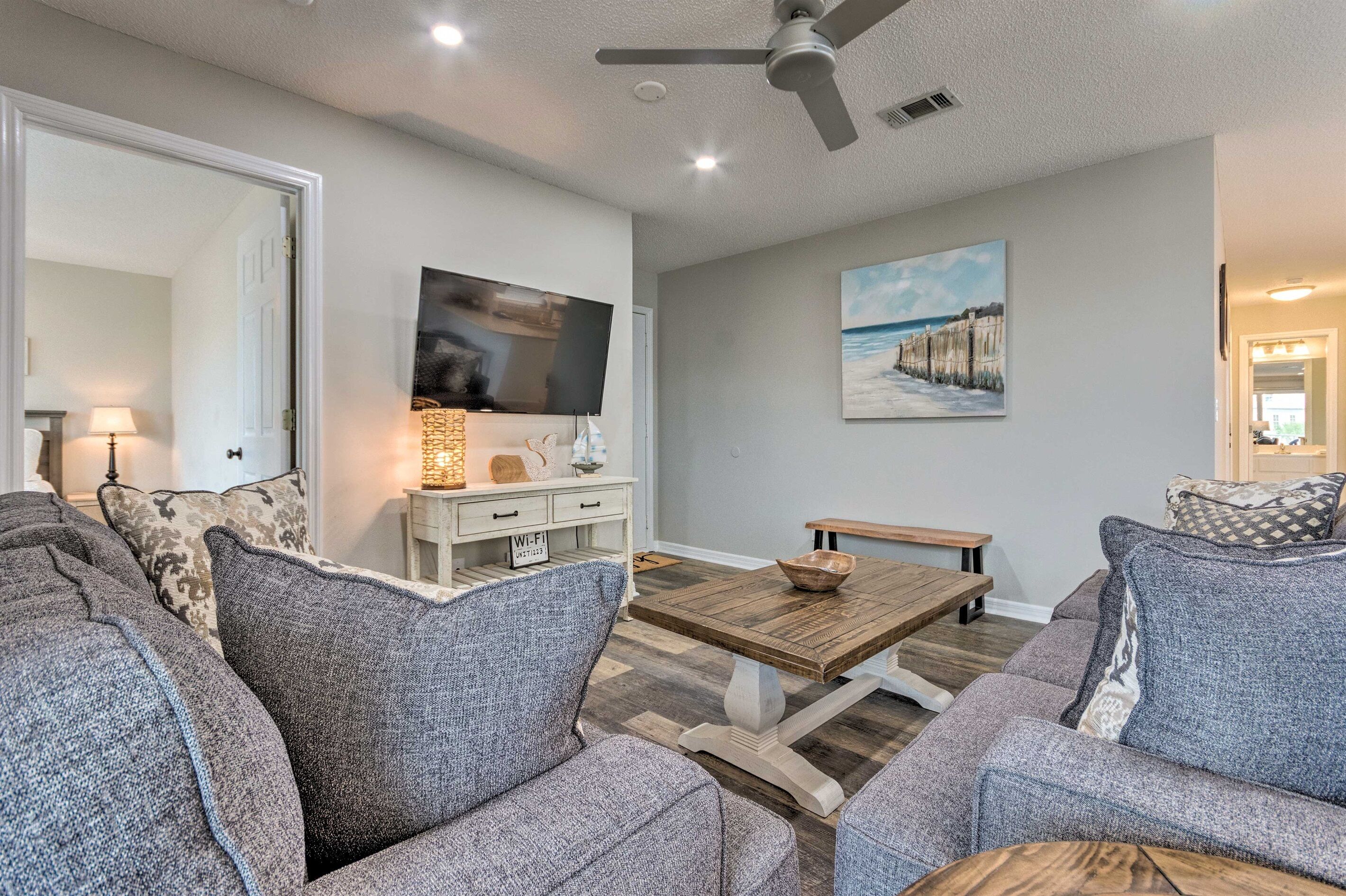 A living room with a sofa set, a wooden coffee table, a flat-screen TV, a painting on the wall, and a ceiling fan.