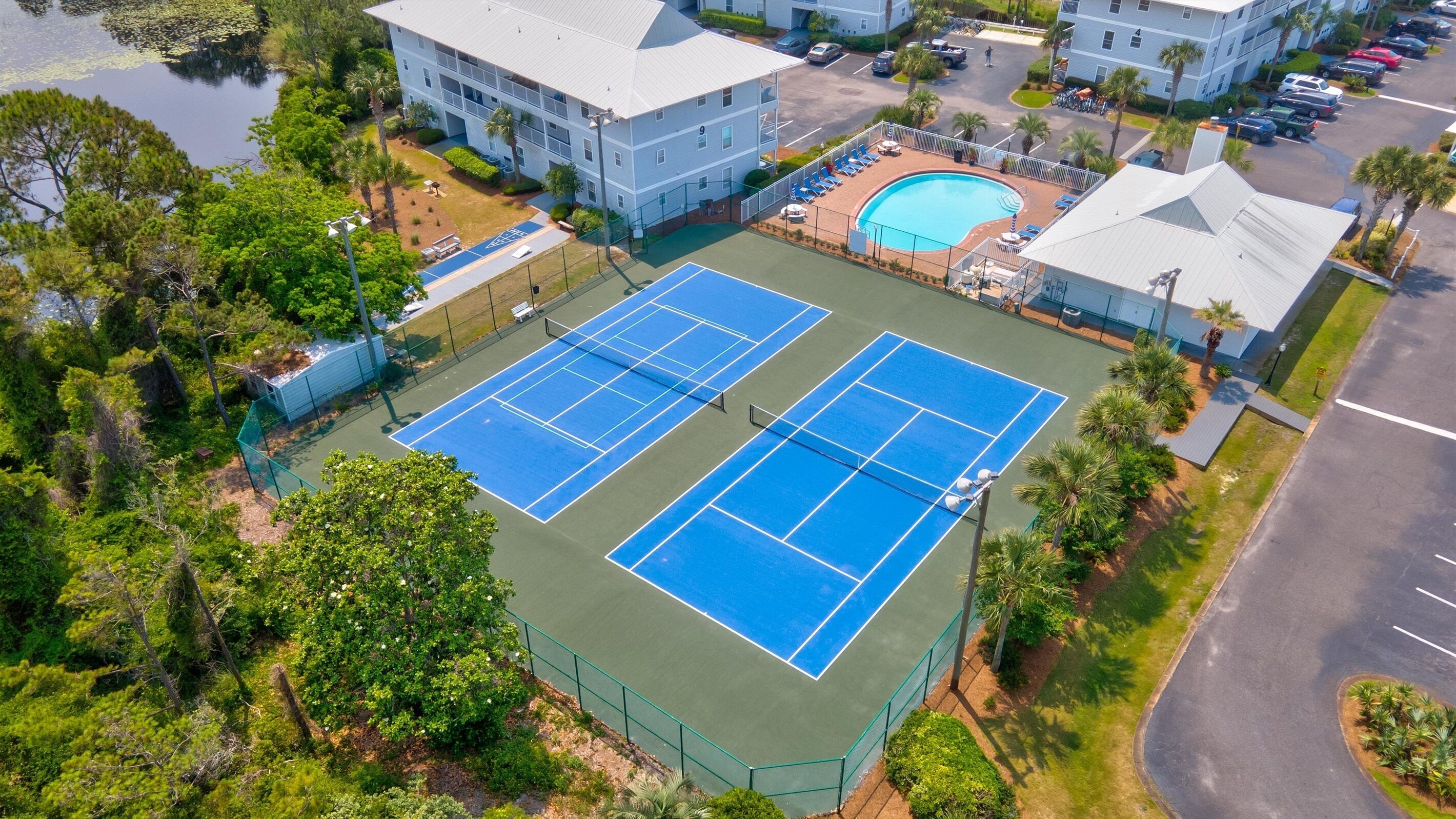An aerial view of a hotel with two tennis courts, a swimming pool, and surrounding greenery.