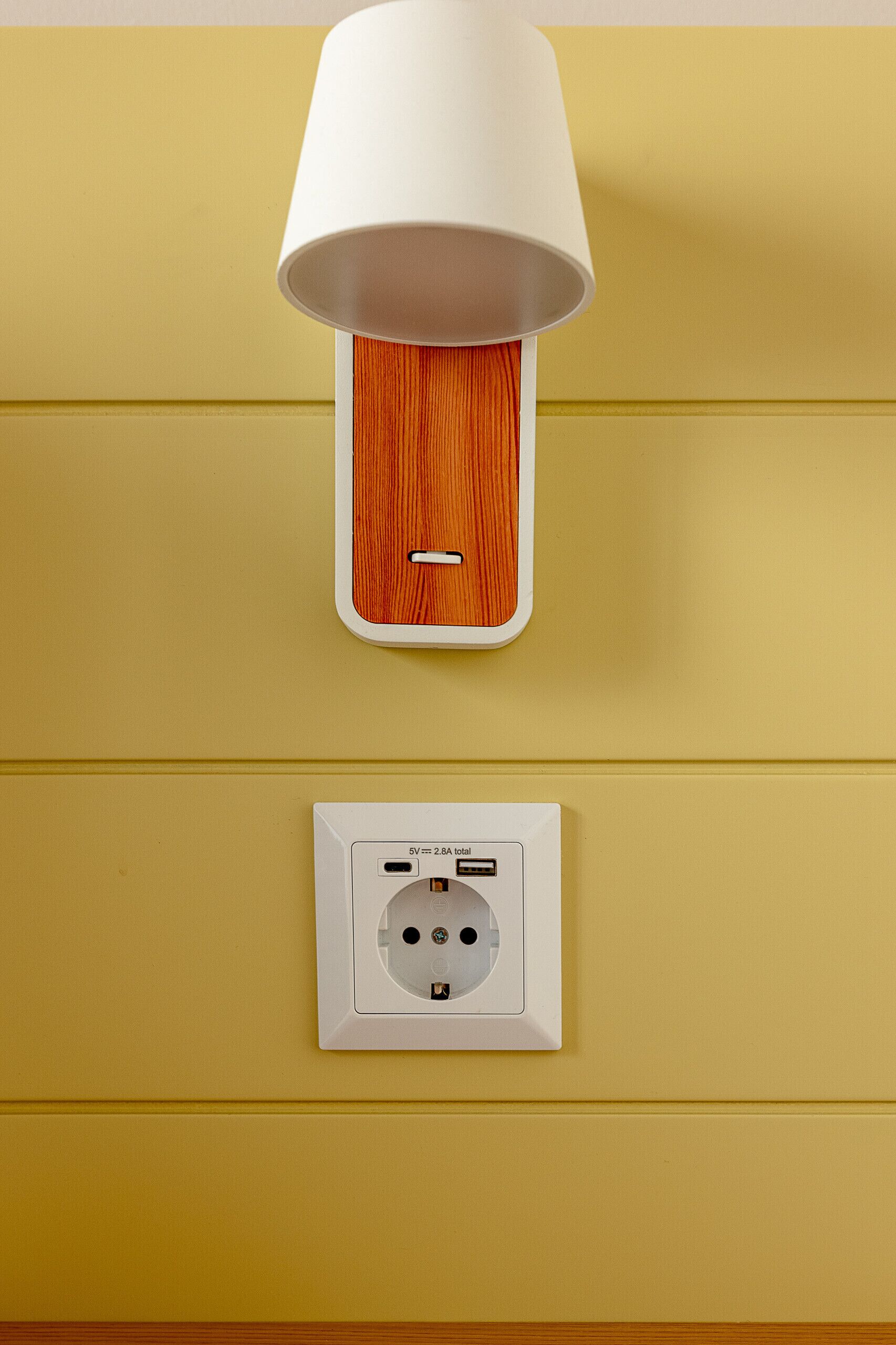 A wall-mounted lamp with a white shade and a wooden base, and a power outlet below it.