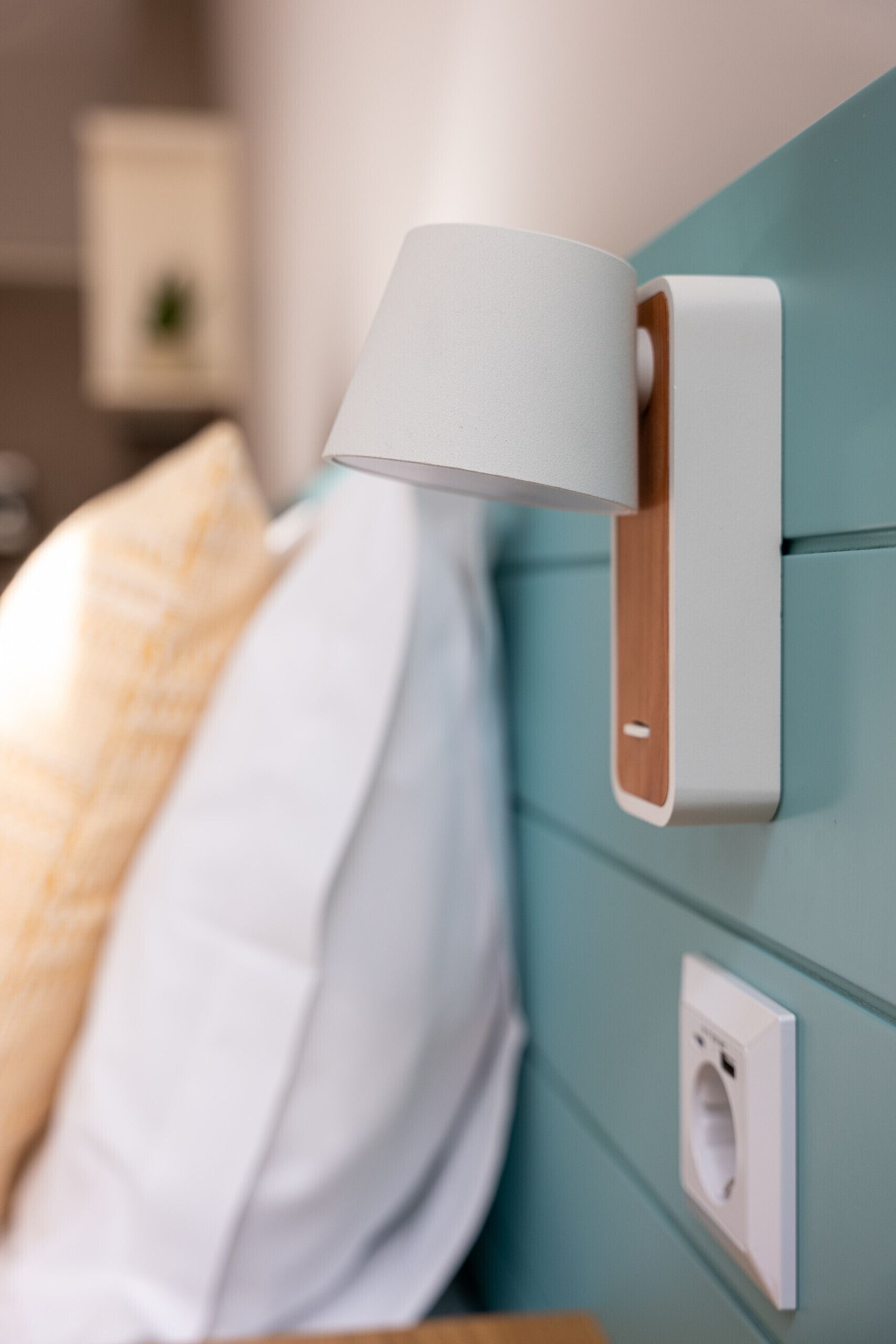 A modern bed with a white pillow and a beige blanket, a wall-mounted lamp, and a power outlet on a teal-colored wall.