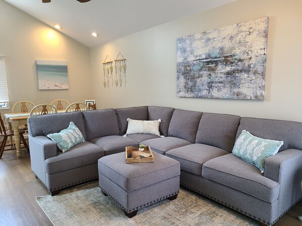 A modern living room with a grey sectional sofa, a small ottoman, and a dining area with wooden chairs and a table.