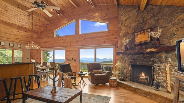 A spacious living room with a stone fireplace, wooden ceiling, and large windows offering a mountain view. - Amazing Views*HotTub*Sauna*GameRooms Dog-friendly! (Beech Mountain)
