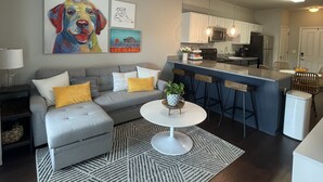 Ein modernes Wohnzimmer mit grauem Sofa, einem runden Couchtisch und einem gemusterten Teppich. Es gibt eine Küchenzeile mit einer Bar und einem Deckenventilator.