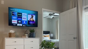 Ein modernes Wohnzimmer mit einem an der Wand montierten Flachbildfernseher, der verschiedene Streaming-Dienste zeigt. Es gibt einen weißen Konsolentisch mit Schubladen, einen runden weißen Tisch mit einer Zierpflanze und einen gemusterten Teppich auf dem Boden. Im Hintergrund ist eine Tür mit einem Deckenventilator zu sehen.