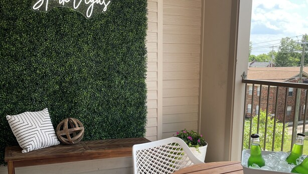 Ein Balkon mit grüner Wand, einer Holzbank, Korbstühlen, einem Holztisch und einem weißen Korb mit grünen Flaschen.