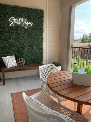 Un balcon agrémenté d’un mur végétal, d’un banc en bois, de chaises en osier, d’une table en bois et d’un panier blanc contenant des bouteilles vertes.