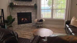 Un salon doté d’une cheminée en pierre, d’un téléviseur à écran plat fixé au-dessus, d’une grande fenêtre avec des stores et d’un canapé en cuir.