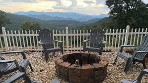 Un foyer circulaire, entouré de six chaises Adirondack, est installé sur une surface de gravier. Une clôture en bois peinte en blanc et un paysage montagneux complètent le décor.