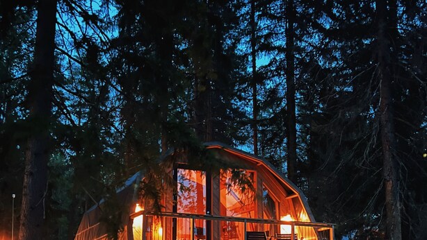 Una cabaña de madera con ventanas amplias y una terraza, rodeada de un bosque tupido.