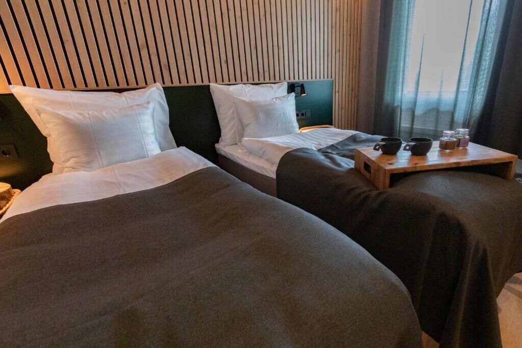 A hotel room with two beds, a wooden headboard, a bedside table with cups and a tray, and a window with curtains.