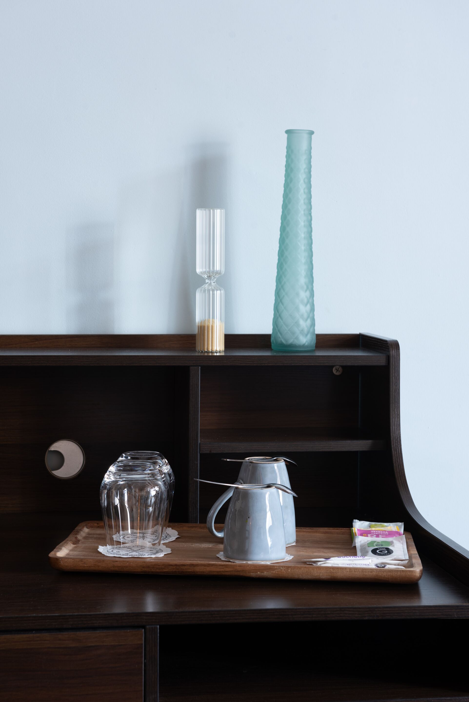 A wooden tray with a glass pitcher, a clear glass, and a teal vase on a dark wooden shelf.