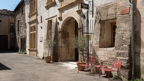 A stone building with a sign reading "Place de l'Anniversaire." There are two red metal chairs and a table outside, with potted plants nearby. - La Maison des Seigneurs (Cucuron)