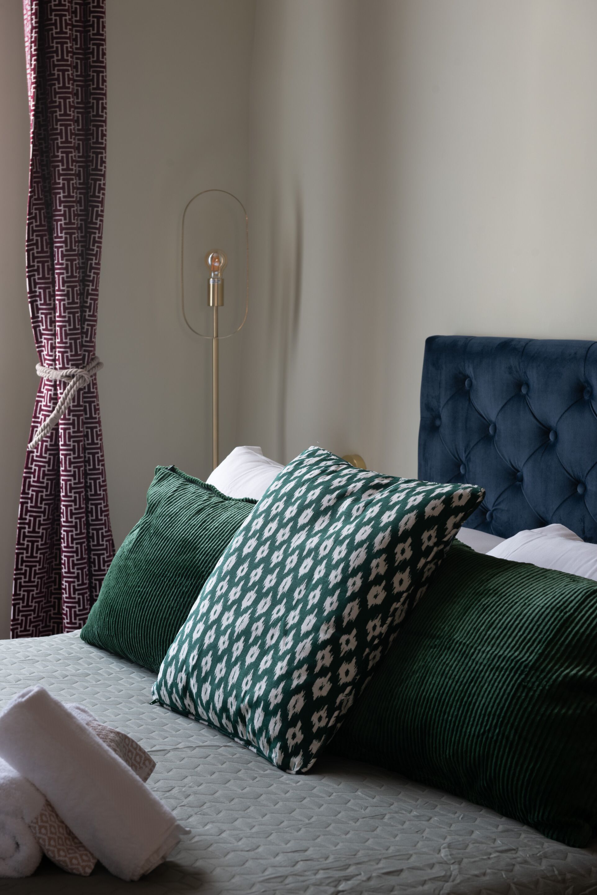 Cama bem arrumada com almofadas de padrão verde e branco, cabeceira estofada e abajur em quarto de hotel.