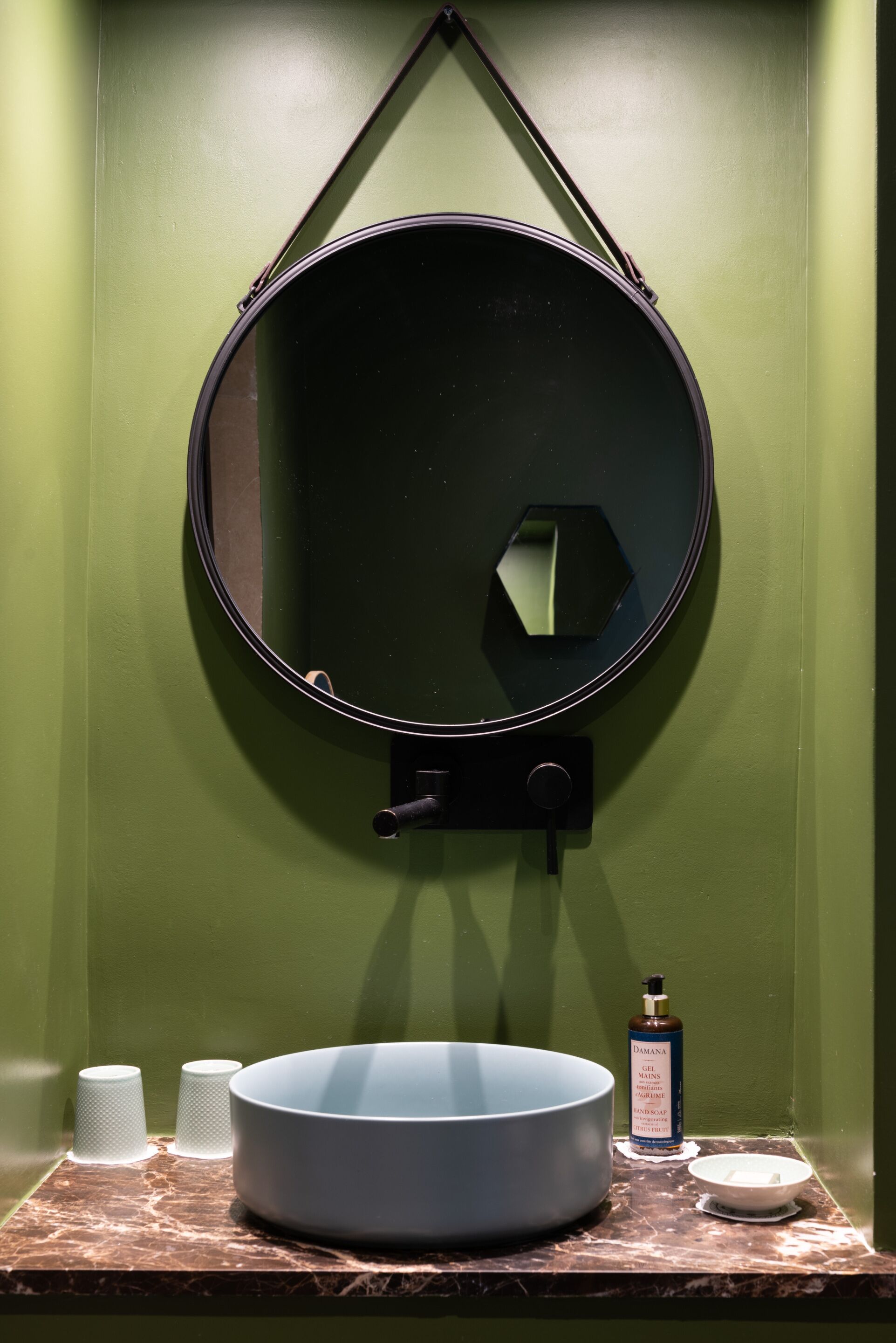 A bathroom with a large round mirror, a bowl sink, and a bottle of hand soap on a marble countertop.