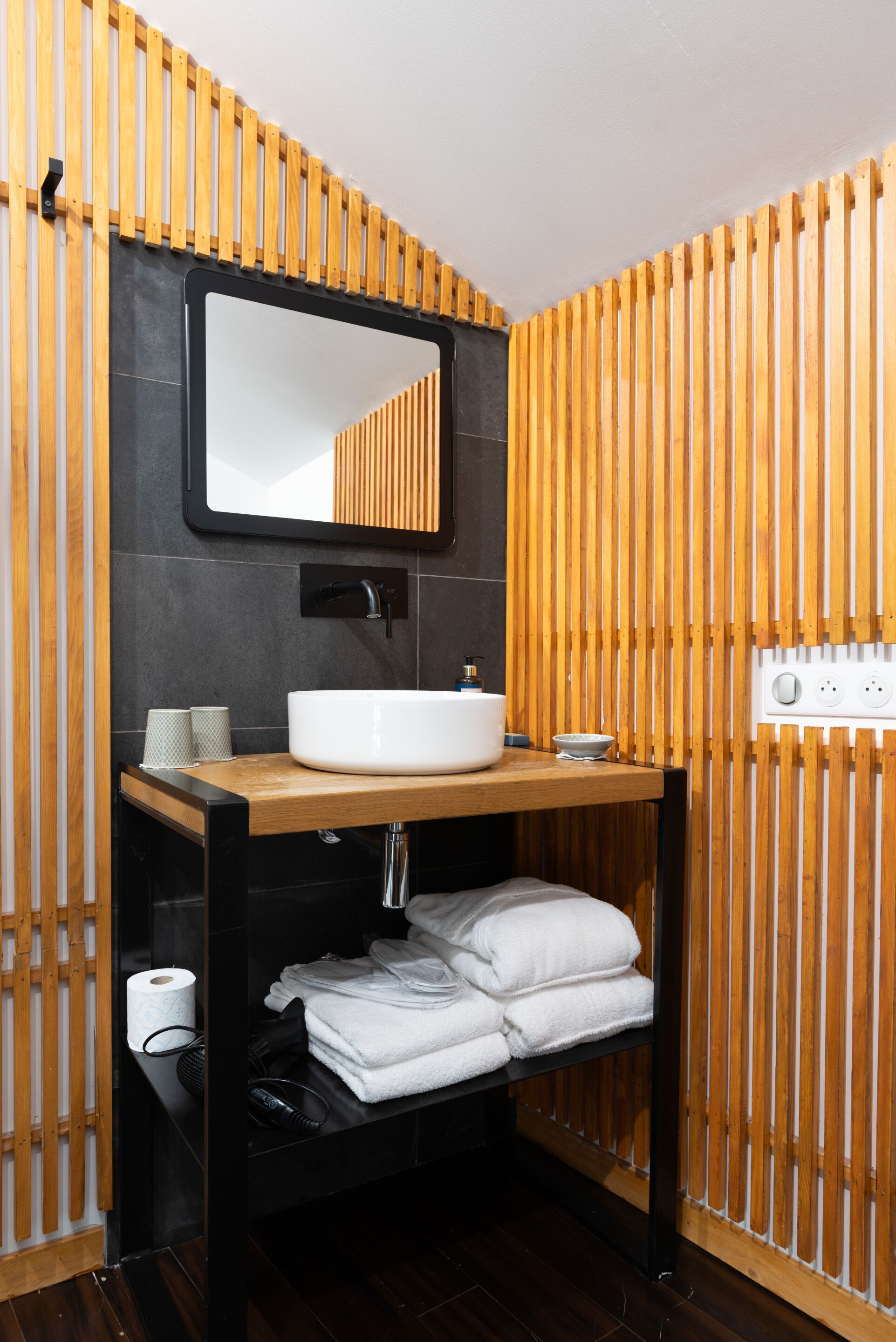 Un baño moderno con pared de listones de madera, un espejo grande, un lavabo blanco y toallas dobladas con cuidado.