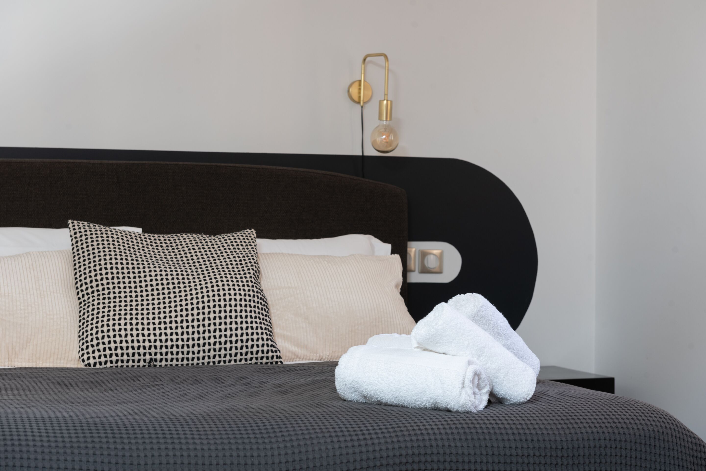Een netjes opgemaakt bed met een donker hoofdbord, een gestreept kussen en een opgevouwen witte handdoek.