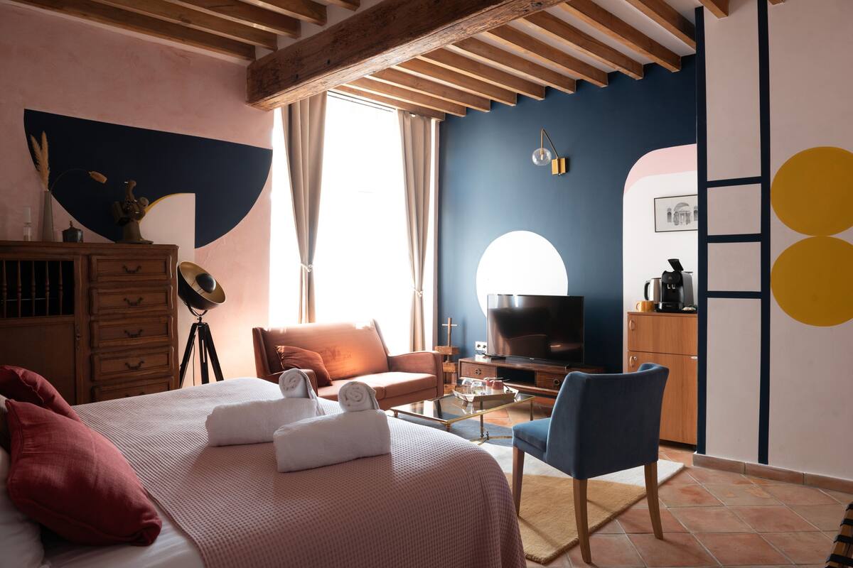 A hotel room with a bed, a desk, a chair, a TV, and a kitchenette.