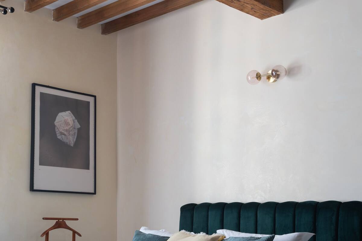 A bedroom with a bed, wooden beams, a framed picture, and a small wooden stool.
