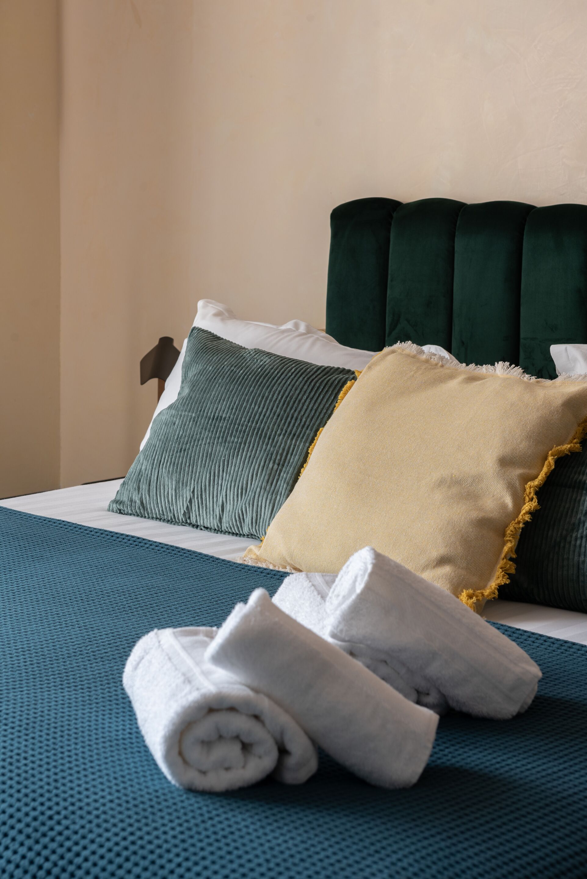 Een netjes opgemaakt bed met een blauwgroene deken, twee kussens (een groen en een beige) en een opgerolde witte handdoek op een blauwe, geribbelde mat.