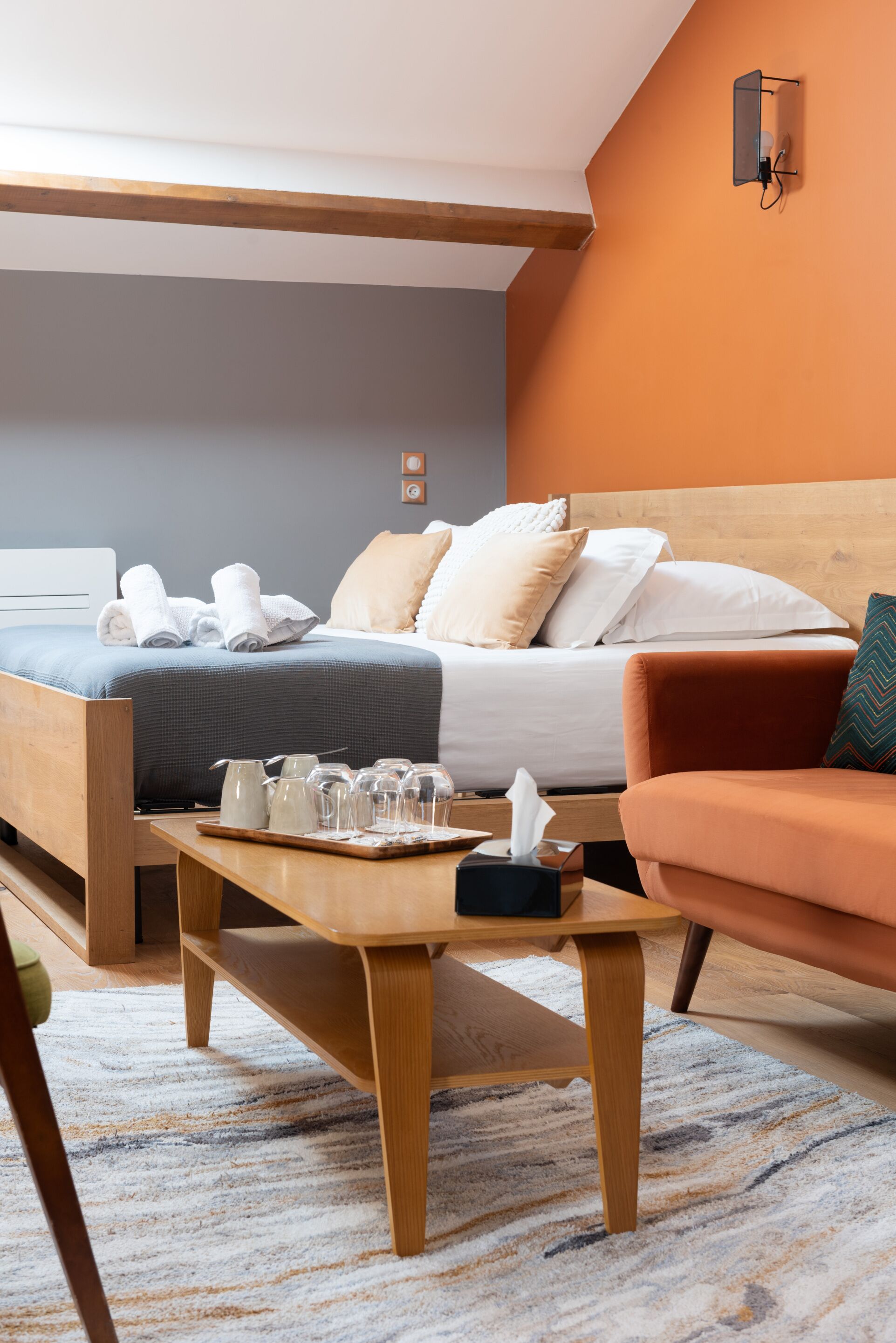 Quarto de hotel com cama, mesa de centro de madeira, sofá e luminária na parede.
