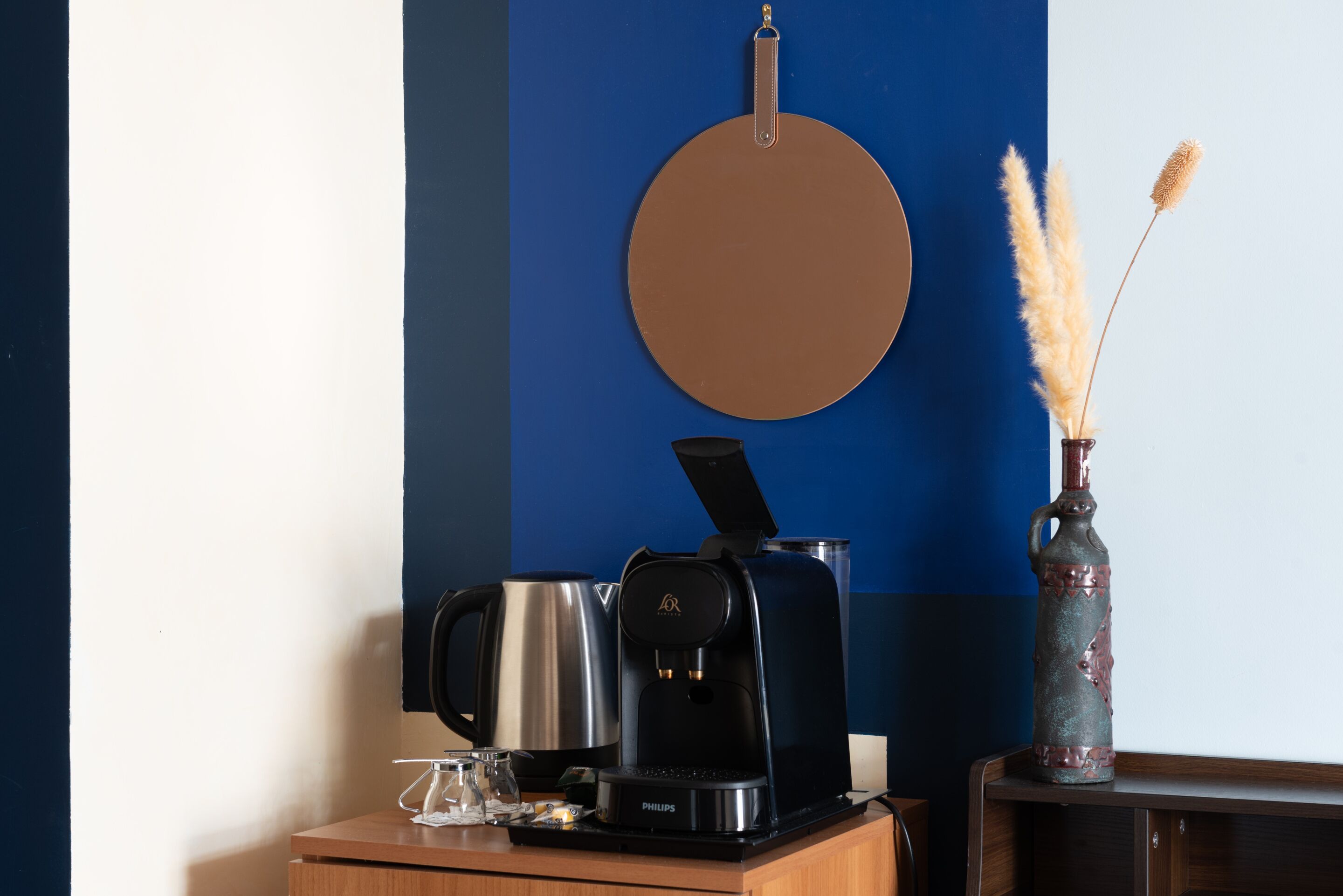 A coffee maker, a vase with pampas grass, and a round mirror on a wooden surface.