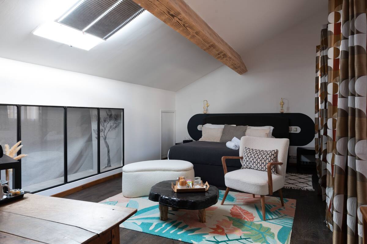 A modern bedroom with a large bed, a wooden ceiling beam, a black headboard, a white armchair, a round ottoman, and a large window with a view of trees.
