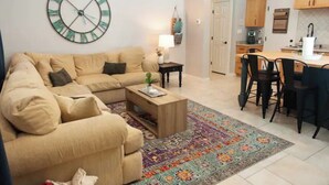 A living room with a beige sofa, a coffee table, and a colorful rug. There is a kitchen area with wooden cabinets and a dining table with chairs.