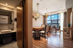 Una habitación de hotel moderna con suelo de madera, una zona de comedor con mesa y sillas, un ventanal grande y un baño con lavamanos y espejo.
