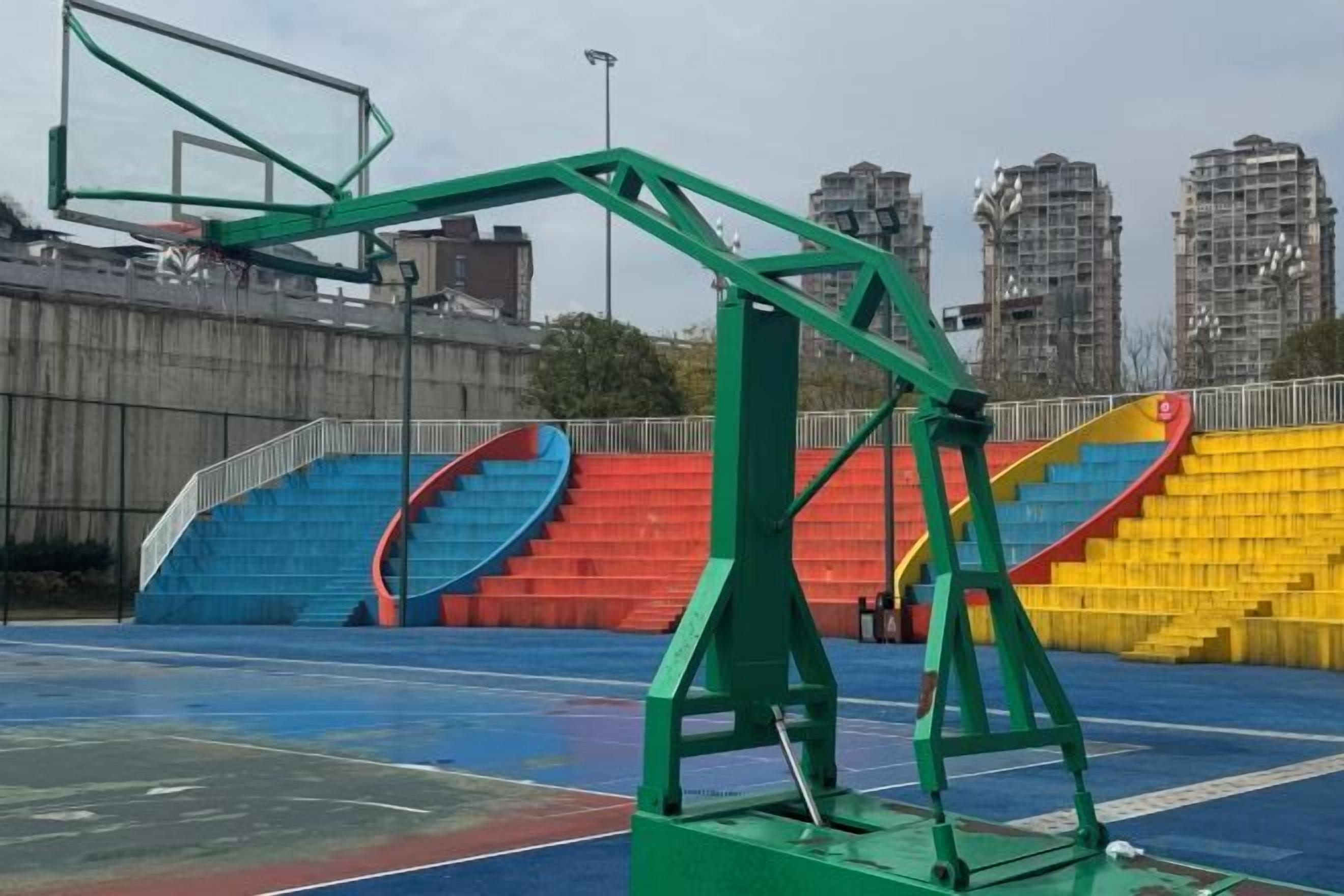 Een buitenbasketbalveld met een groene basketring, blauwe en rode tribunes en een stadsgezicht op de achtergrond.