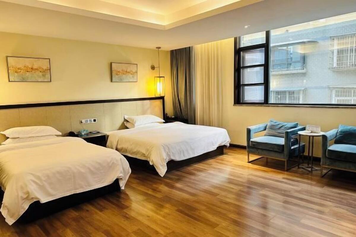 Habitación de hotel con dos camas, una mesita, dos sillas, un ventanal con vistas y obras de arte en las paredes.