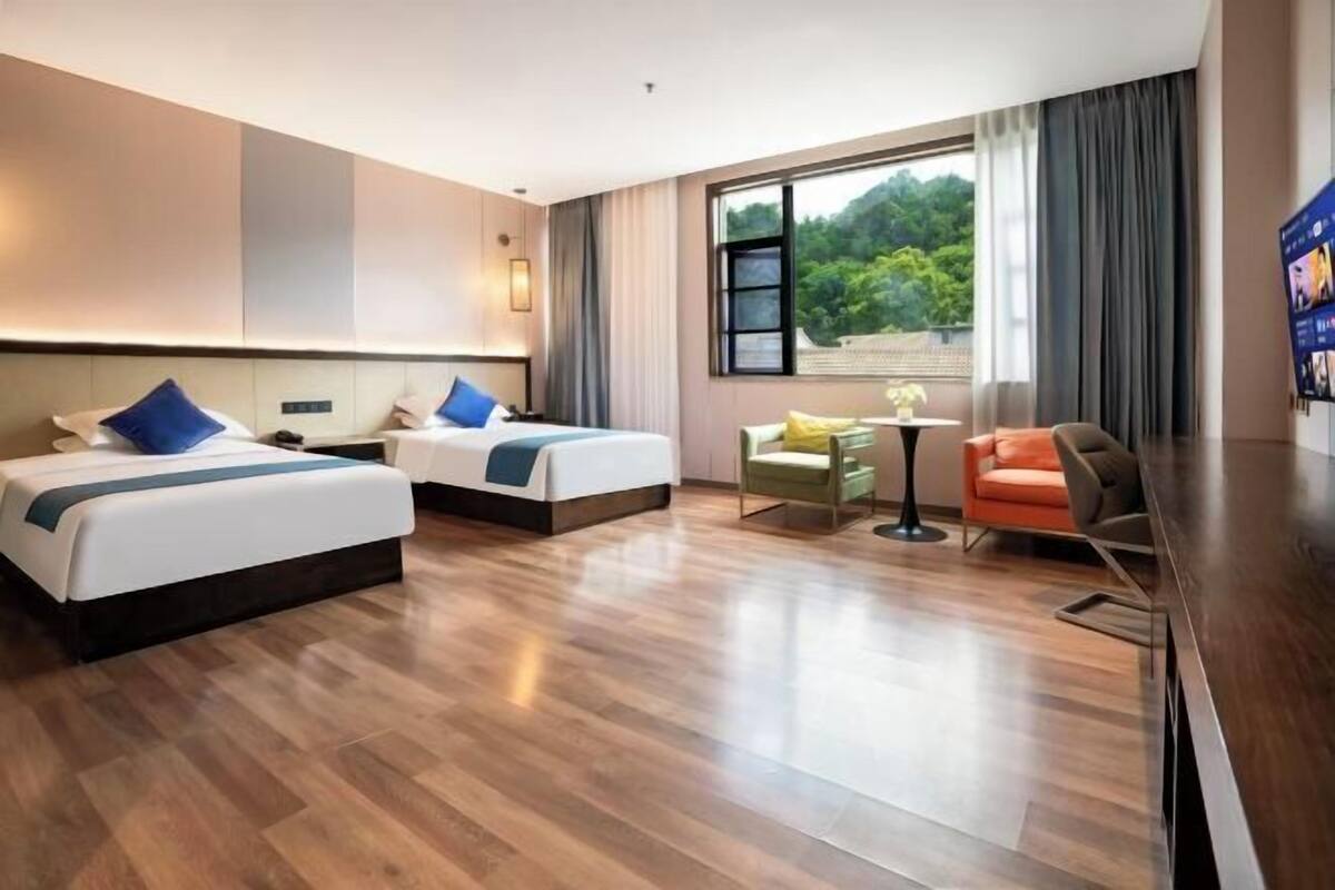 Habitación de hotel con dos camas, una mesita, una silla y un ventanal con vistas a zonas verdes.