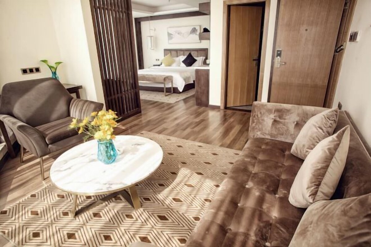 A modern hotel room with a brown leather sofa, a round coffee table with a vase, and a patterned rug.