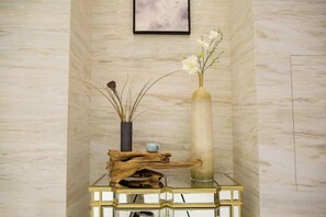Una pared de mármol con un cuadro enmarcado, una mesa de vidrio con un florero y ramas secas, y un florero con flores.