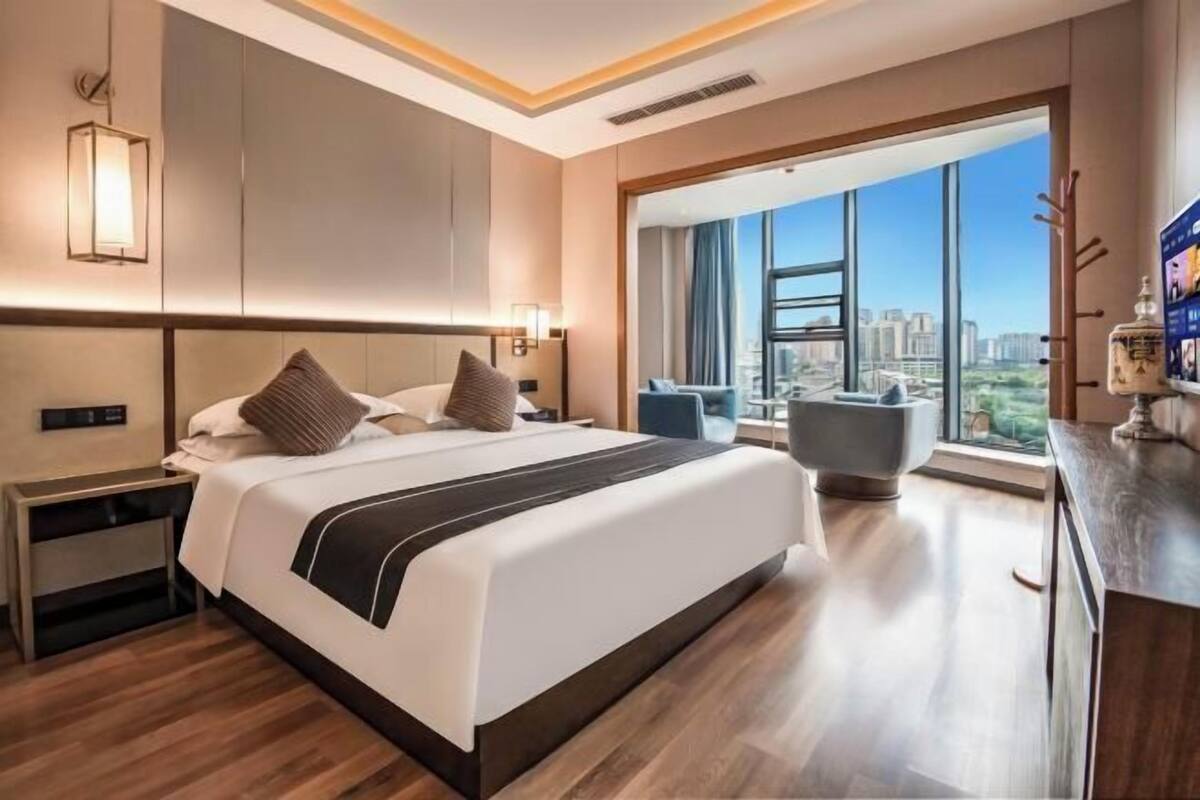 Una habitación de hotel moderna con una cama grande, una mesita de noche, un escritorio, una silla y un amplio ventanal con vistas a la ciudad.